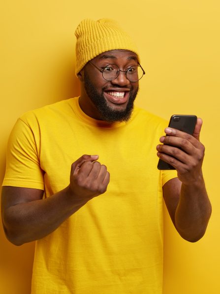 Happy joyous black man clenches fist with triumph, watches football match online, focused in smartphone device, wears round spectacles, yellow clothes, stands indoor, reads funny news on webpage